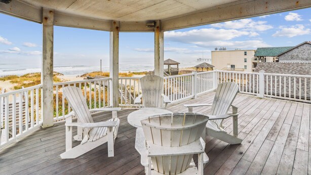 Una terrazza in legno con poltrone Adirondack e un tavolo, con vista su una spiaggia e degli edifici.