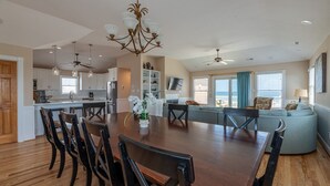 Een ruime woonkamer met een grote houten eettafel, diverse stoelen, een bank en een aan de muur bevestigde televisie.