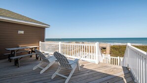 Een houten terras met picknicktafels en stoelen, een witte balustrade en uitzicht op zee.