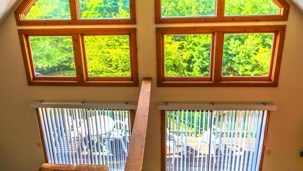 Quarto estilo loft com teto inclinado, grandes janelas com vista para uma floresta, ventilador de teto e um longo banco de madeira.