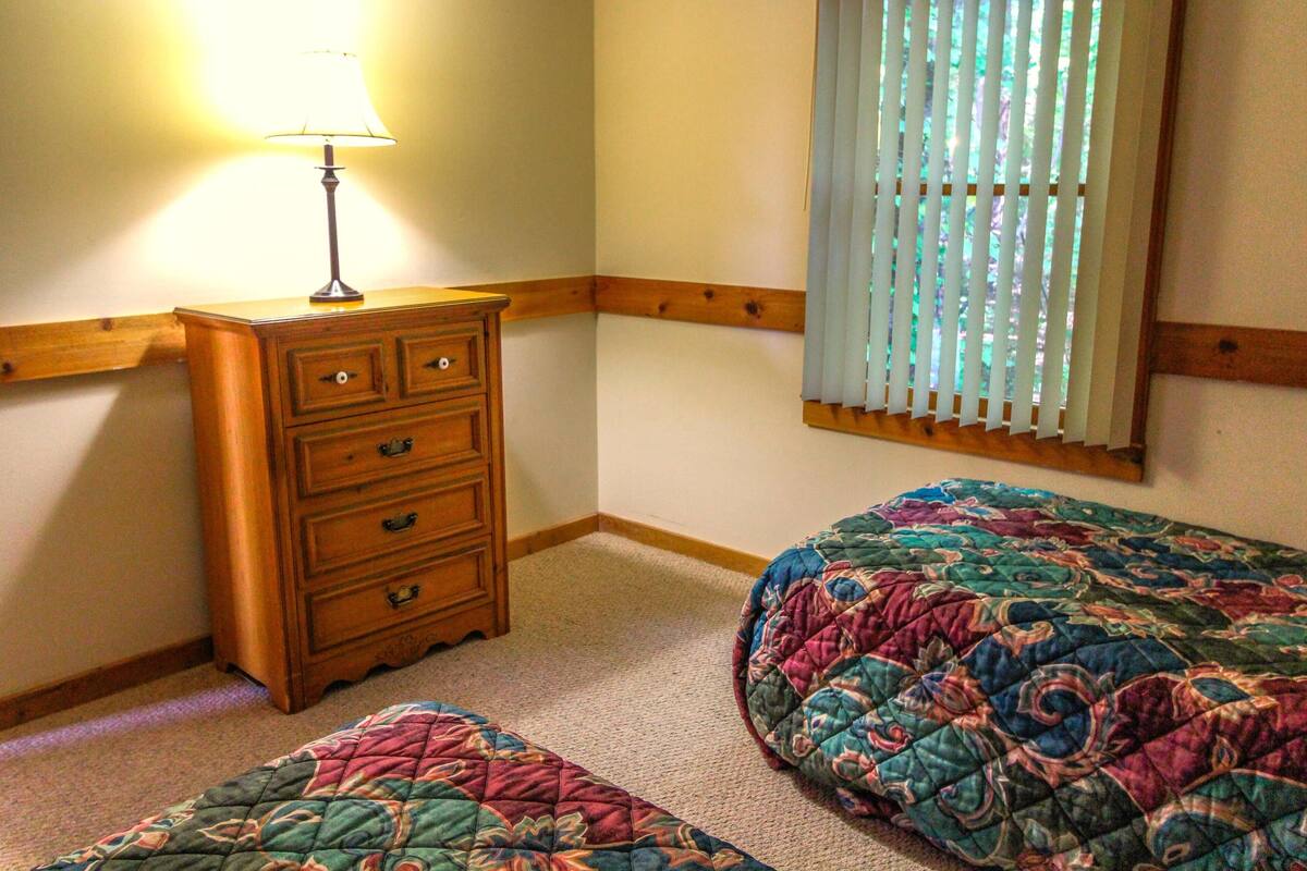 Un dormitorio con un aparador de madera, dos camas con colchas de colores, una ventana con persianas y una lámpara sobre el aparador.