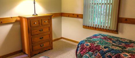 A bedroom with a wooden dresser, two beds with colorful quilts, a window with blinds, and a lamp on the dresser.