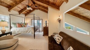 Una camera da letto con un letto a baldacchino, soffitto in legno, vista sul mare attraverso una finestra e un’accogliente zona salotto con un divano marrone e una coperta decorata.