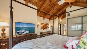 Un dormitorio con una cama grande, un televisor mostrando un barco en el mar, vigas de madera en el techo y vista al mar a través de una ventana.