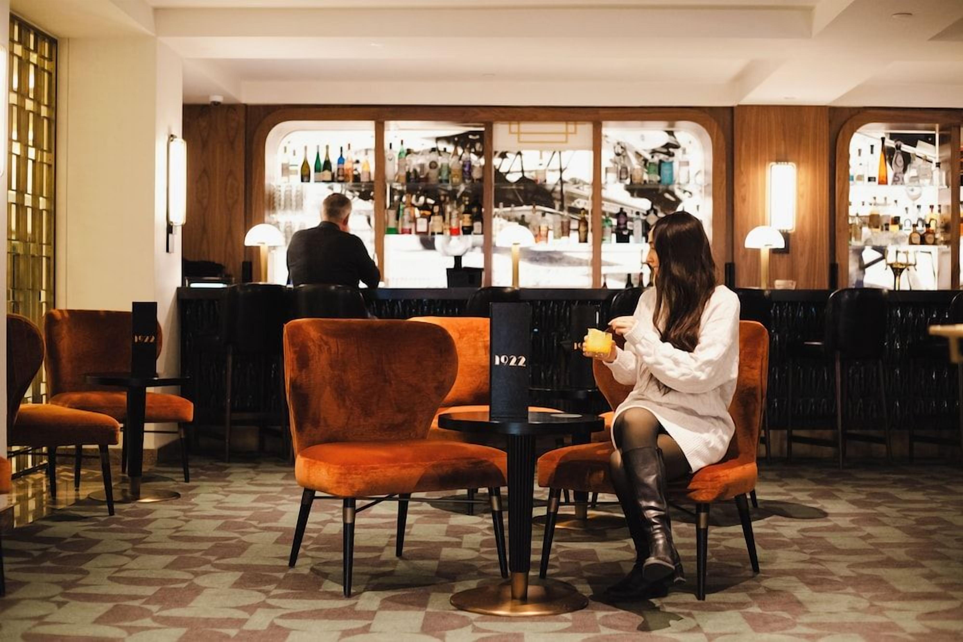 A bar area with a woman seated at a table, a bartender in the background, and a bar counter with bottles.
