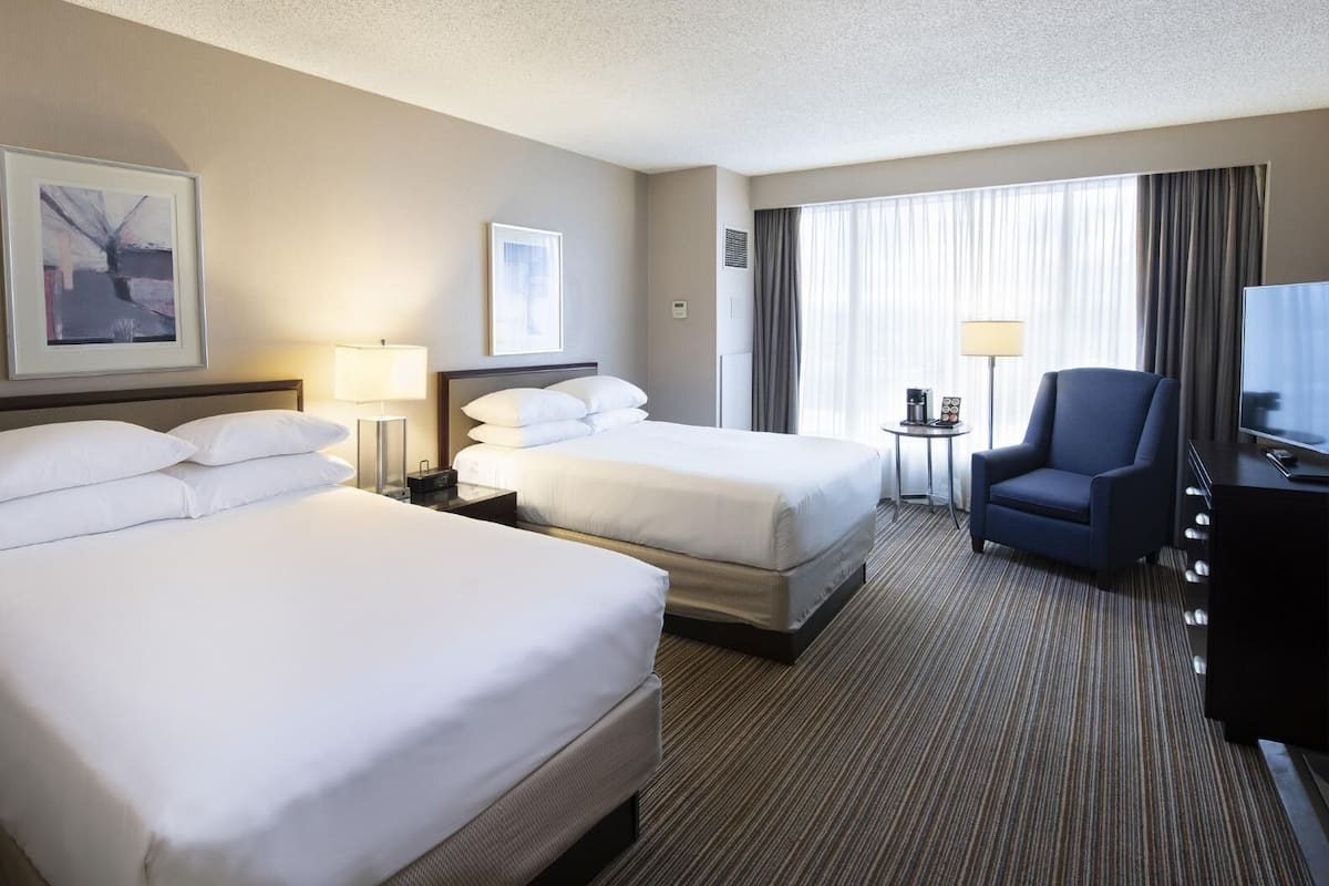 A hotel room with two beds, a desk, a chair, a TV, and a window with curtains.
