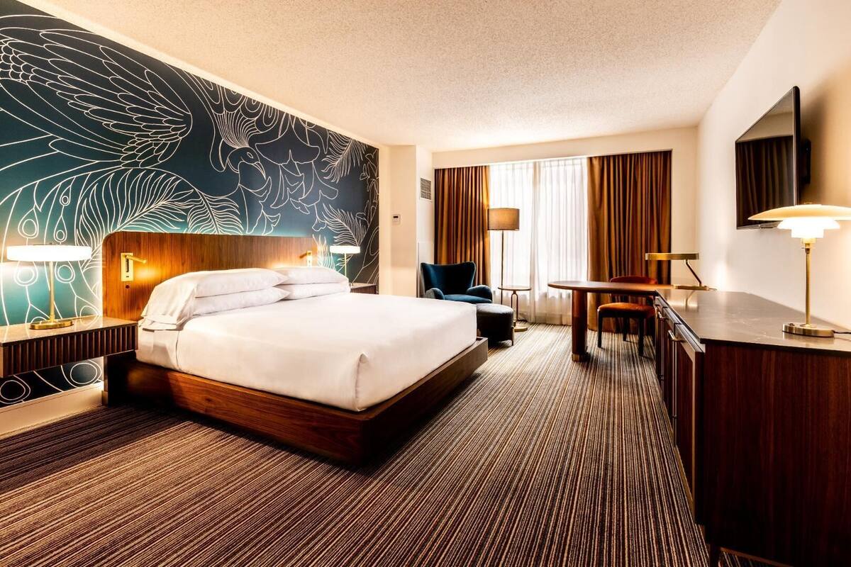 A hotel room with a large bed, a desk, a chair, a TV, and a wall with a leaf pattern.