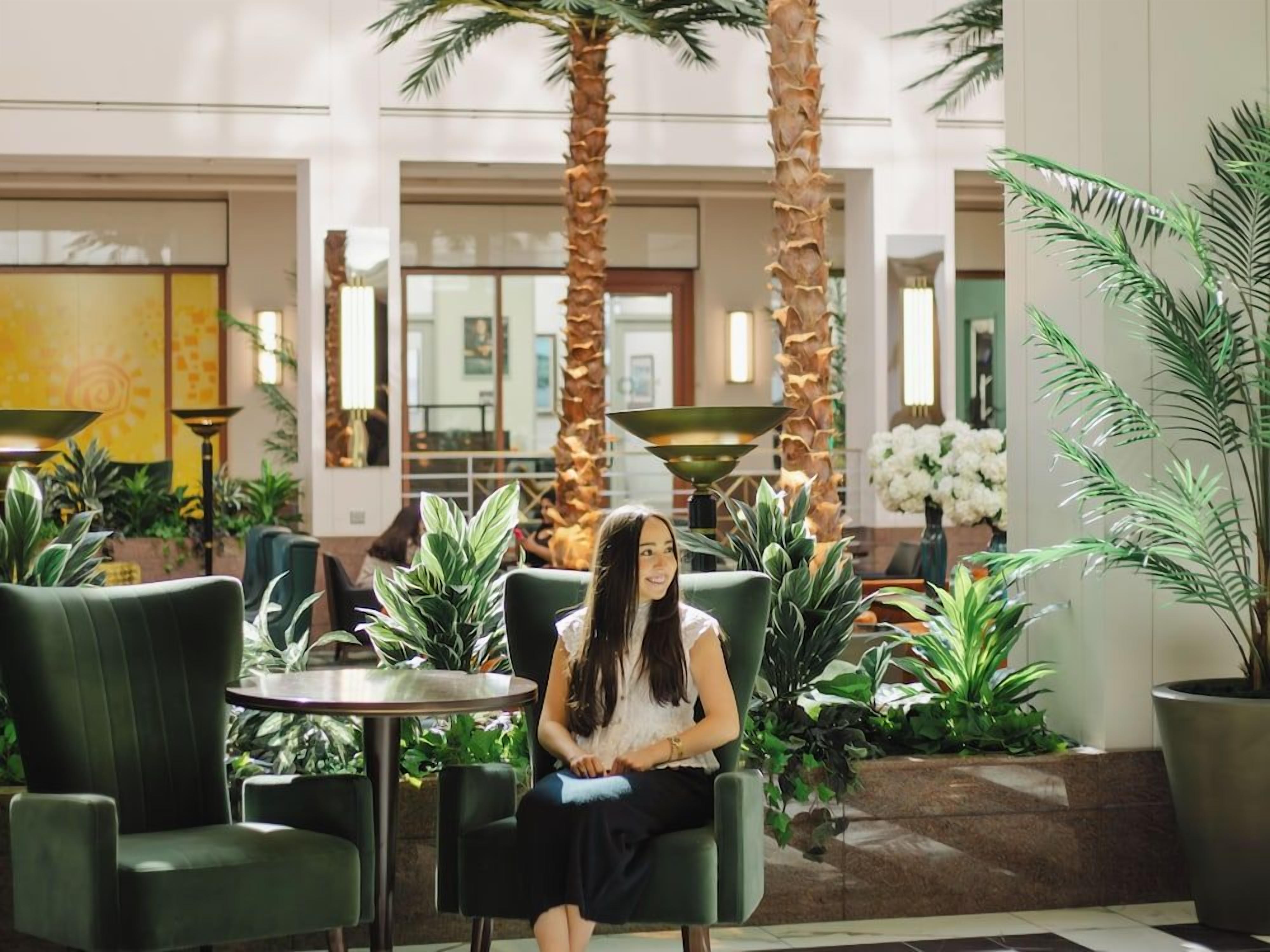 A woman seated in a green chair in a lobby with palm trees and potted plants.