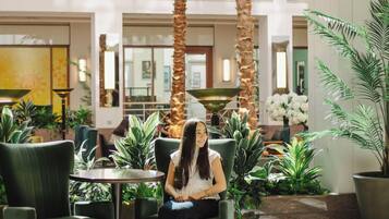 A woman seated in a green chair in a lobby with palm trees and potted plants.