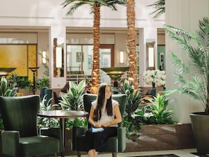 Una mujer sentada en una silla verde en un vestíbulo con palmeras y plantas en macetas.