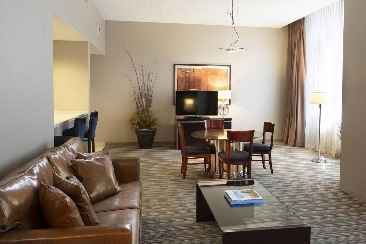 A hotel room with a leather sofa, a dining table with chairs, a flat-screen TV, a potted plant, and a lamp.