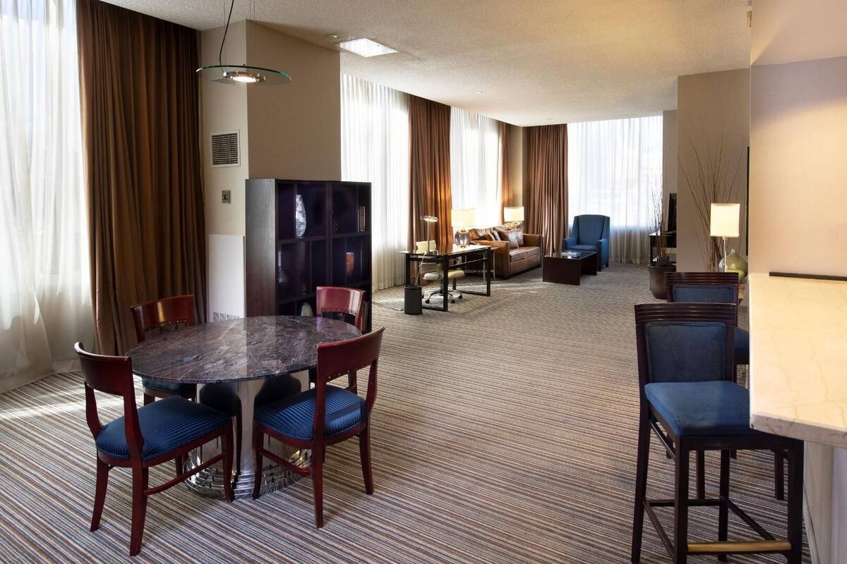 A hotel room with a dining table, chairs, a sofa, a TV, and a lamp.
