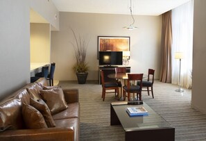 Una habitación de hotel con sofá de cuero, una mesa de comedor con sillas, un televisor de pantalla plana, una planta en maceta y una lámpara.