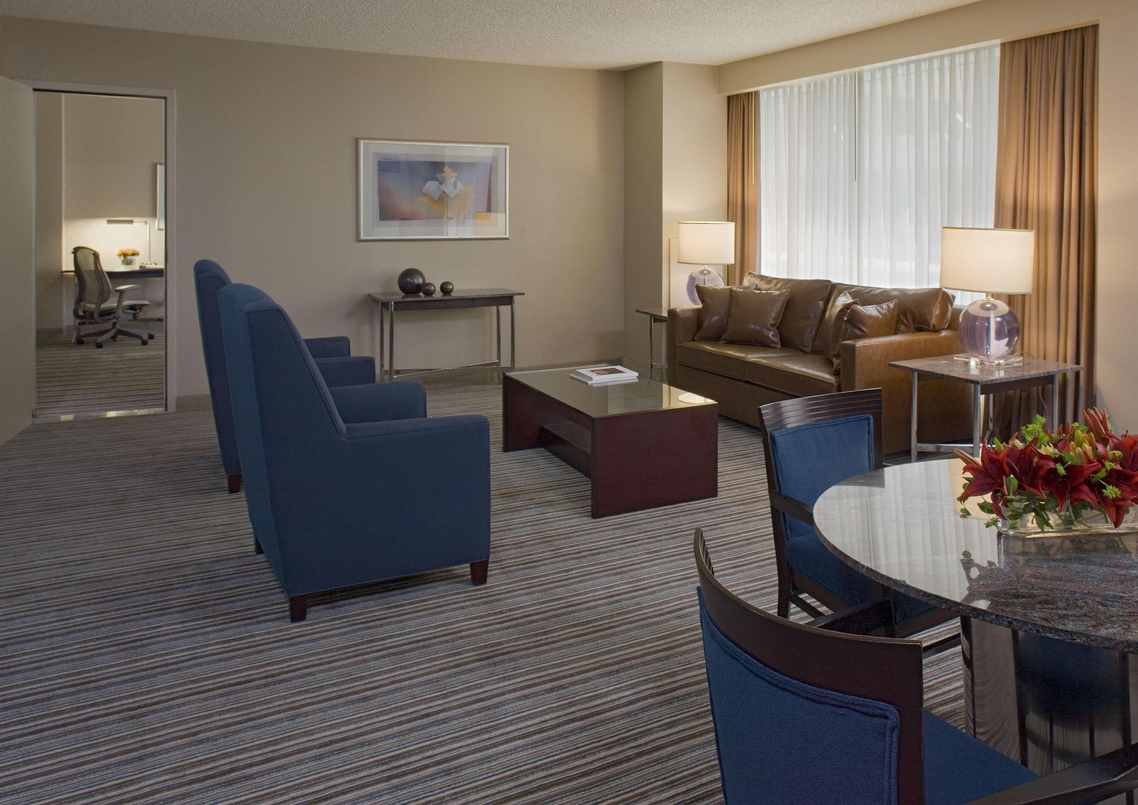 Une chambre d’hôtel comprenant un canapé, deux fauteuils, une table basse, une table à manger avec des chaises, une lampe, un tableau et une fenêtre avec des rideaux.