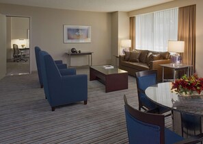 Een hotelkamer met een bank, twee fauteuils, een salontafel, een eettafel met stoelen, een lamp, een schilderij en een raam met gordijnen.