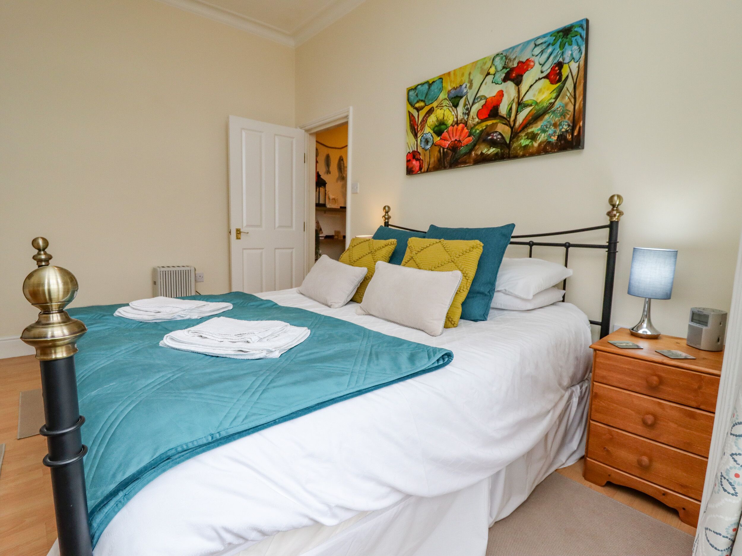 Un lit bien fait, recouvert d’une couvre-lit turquoise, avec des oreillers blancs et un pompon doré sur la tête de lit. Un tableau représentant des fleurs est accroché au mur, ainsi qu’une table de chevet avec une lampe et une petite table en bois.