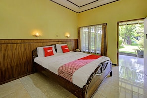 Een hotelkamer met een houten bed, twee rode kussens, een wit-rode dekbedovertrek en uitzicht op een tuin door een groot raam.