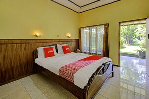 Habitación de hotel con cama de madera, dos almohadas rojas, colcha blanca y roja, y vista a un jardín a través de un gran ventanal.