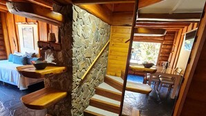 A wooden staircase with a stone wall, a bed with blue bedding, and a dining area with chairs and a table. - PUERTO LACAR LODGE SMARG (San Martín de los Andes)