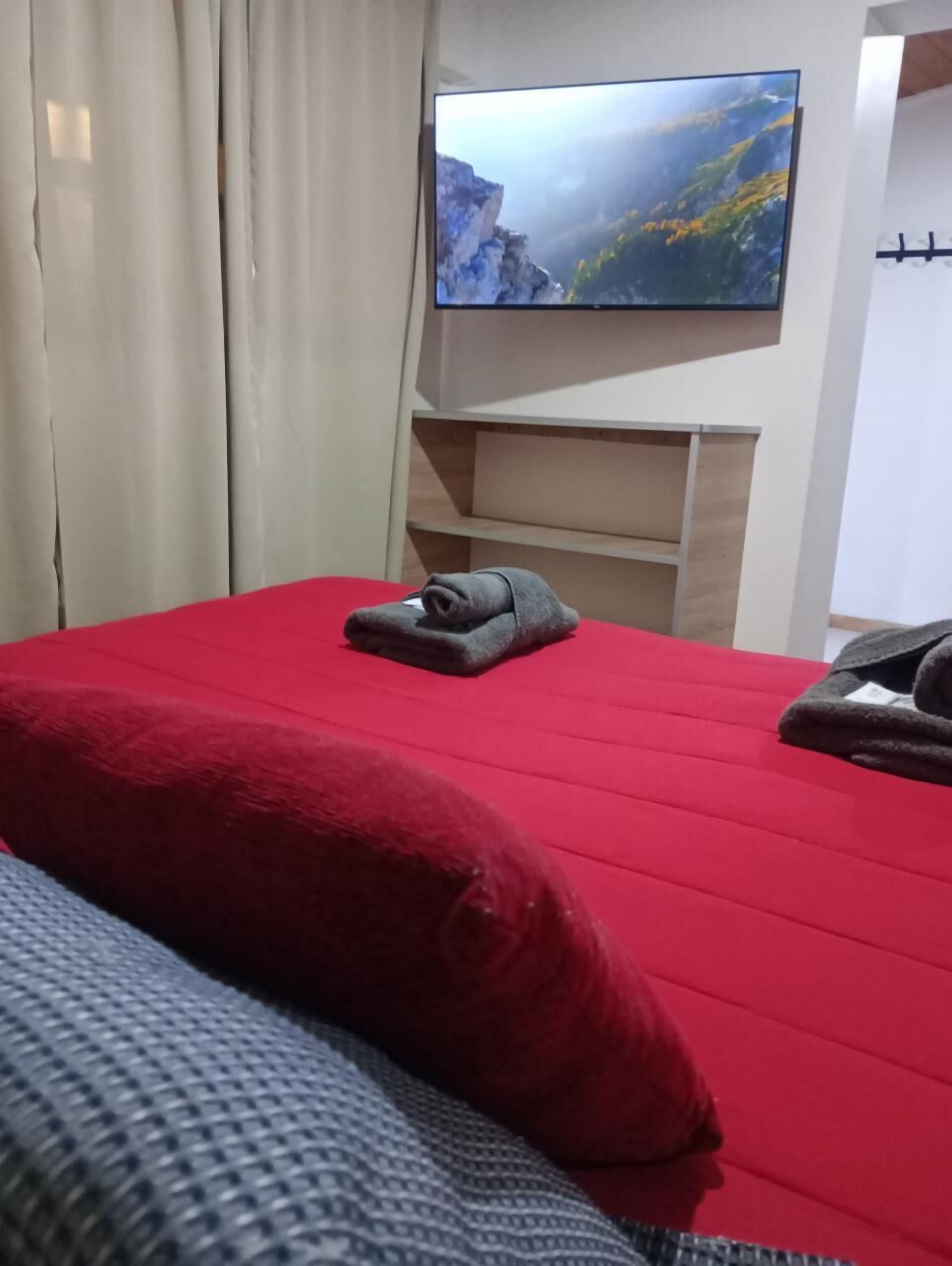 Habitación de hotel con una colcha roja, una manta gris y un televisor en la pared mostrando un paisaje montañoso.