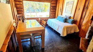 A log cabin room with a wooden dining table, a bed with a white bedspread, and a window with a view of trees. - PUERTO LACAR LODGE SMARG (San Martín de los Andes)