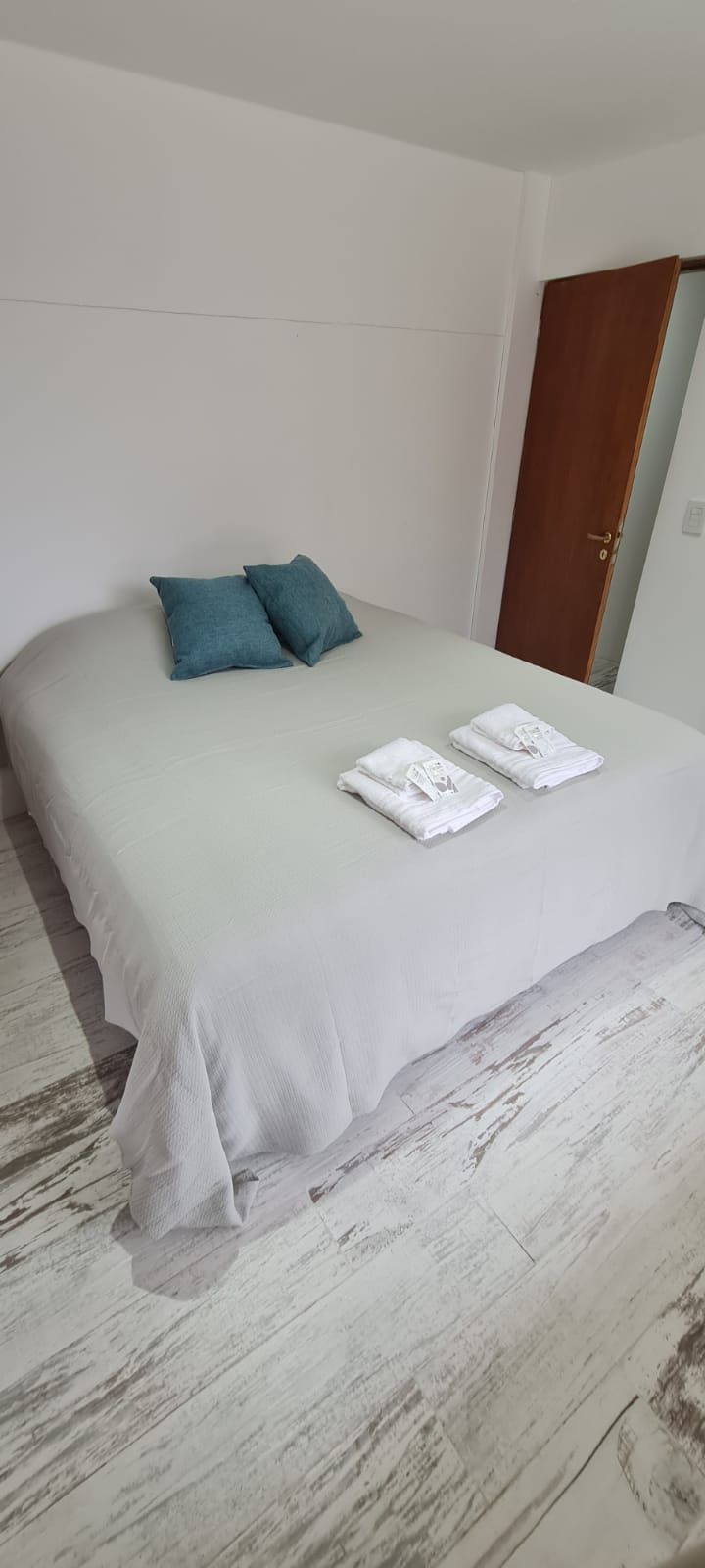 Una cama con una colcha blanca, dos almohadas y toallas dobladas sobre un piso de madera.