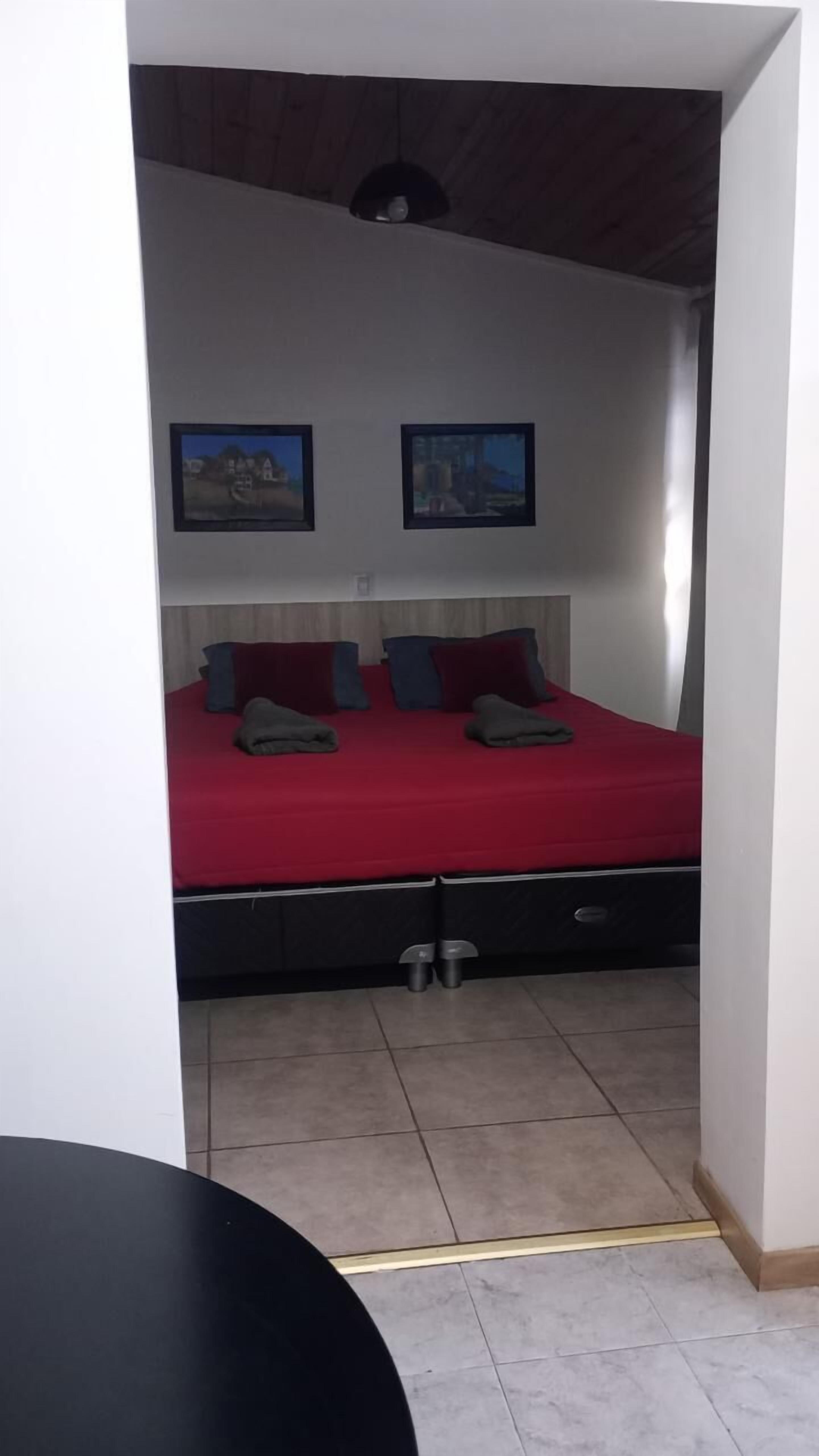A bedroom with a red bedspread, two pillows, and a headboard. There are two framed pictures on the wall.
