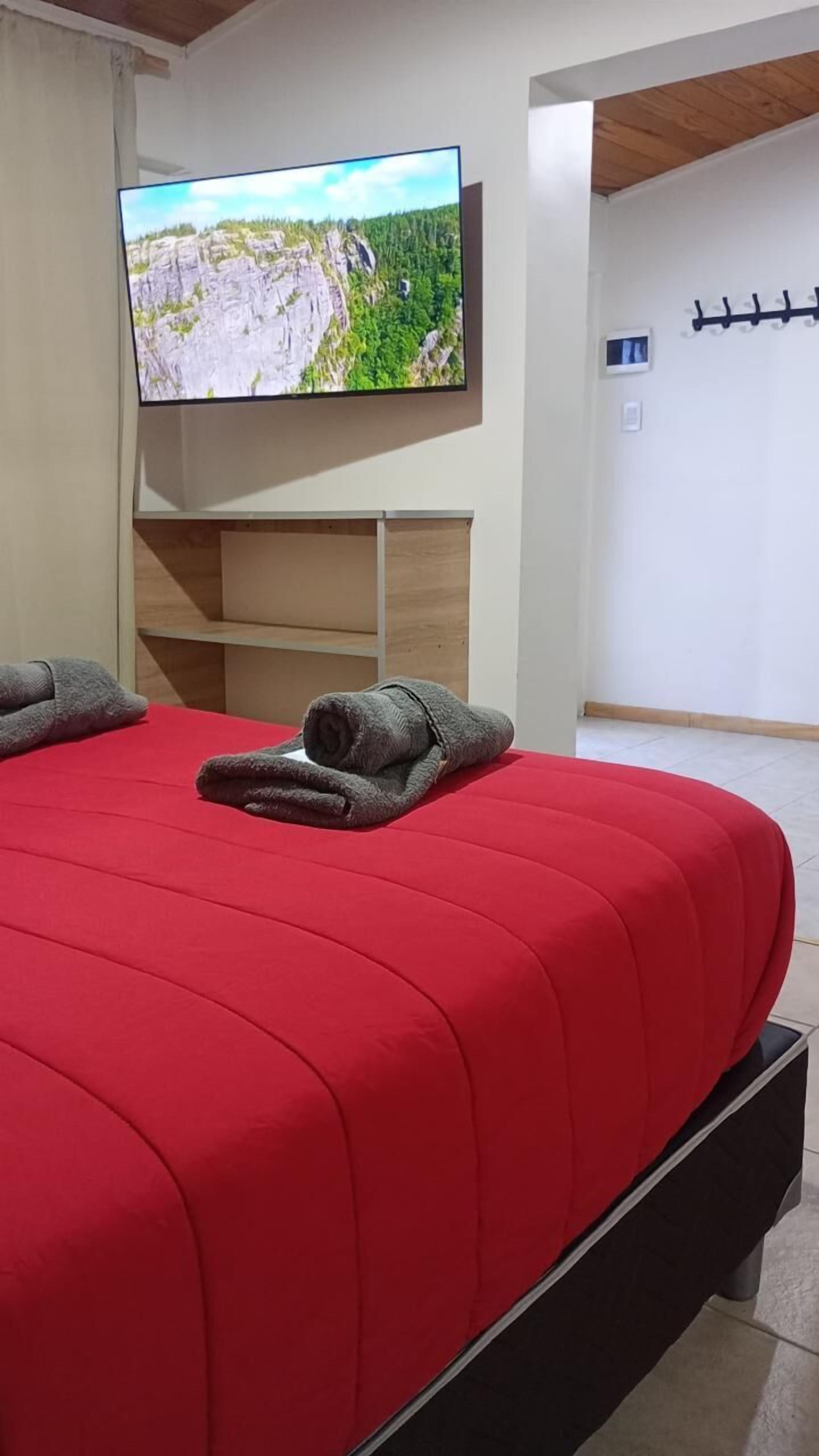 A bedroom with a red bedspread, a television mounted on the wall, and a coat rack.