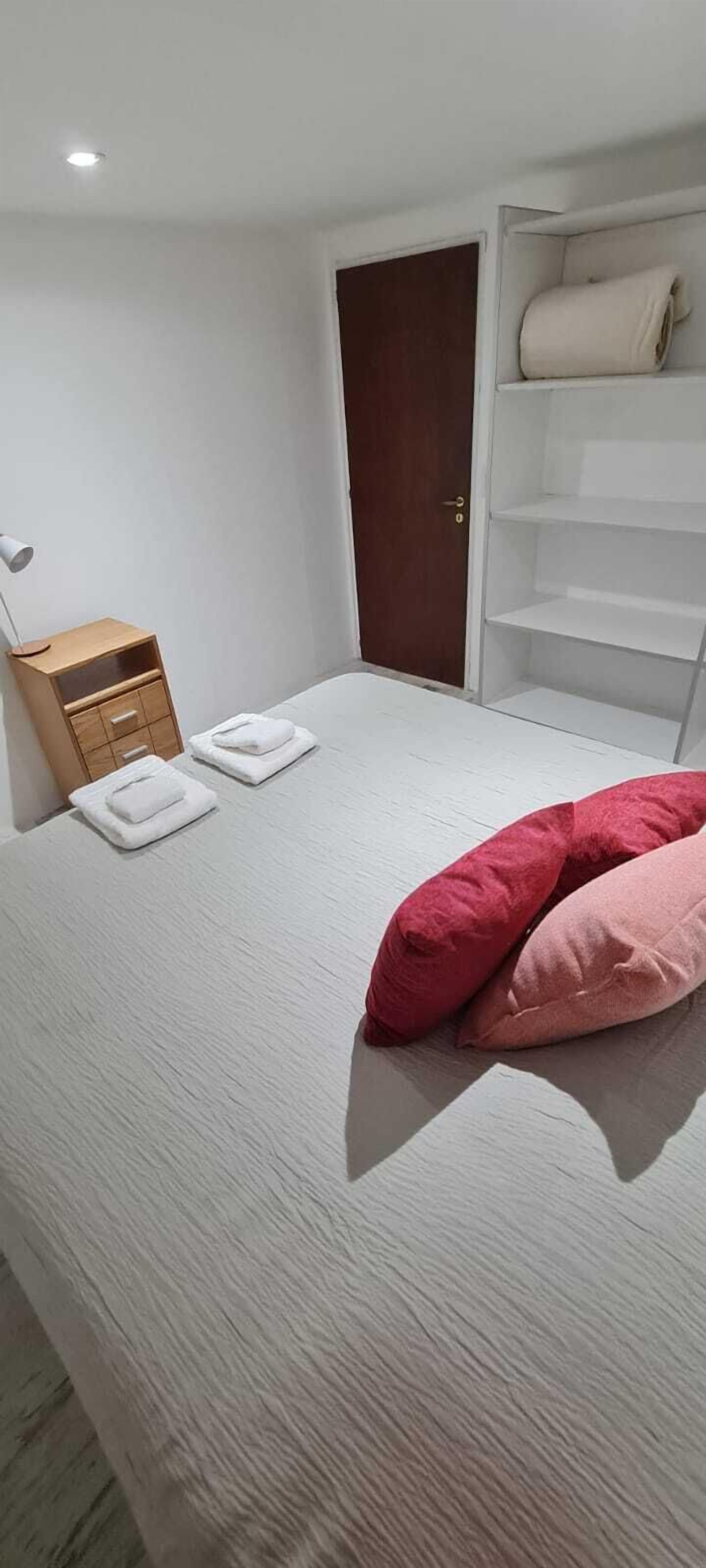 Quarto com cama, mesa de cabeceira, luminária, porta e estante branca.