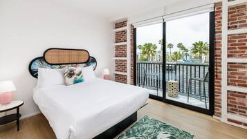 A bedroom with a large bed, a nightstand, a lamp, and a view of a balcony with palm trees.