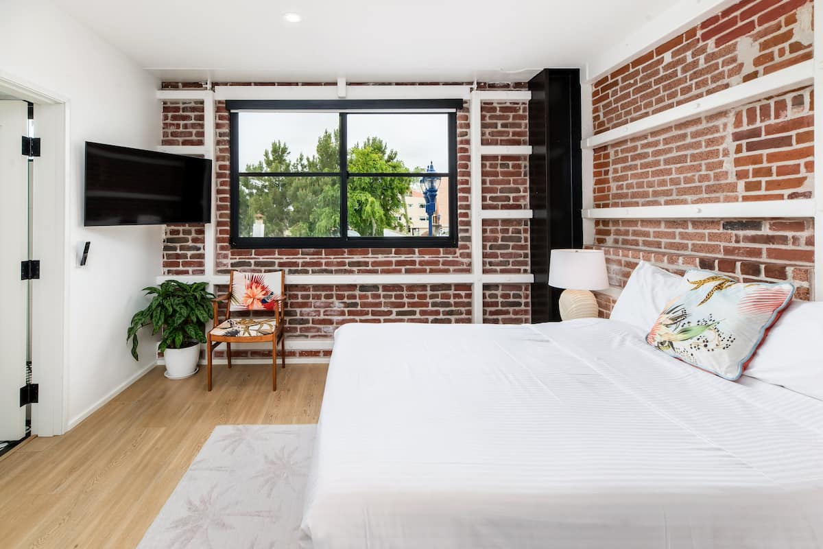 A modern bedroom with a brick wall, a large window, a flat-screen TV, a bedside table with a lamp, a potted plant, and a bed with decorative pillows.