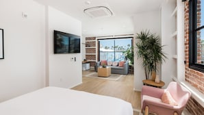 Ein modernes Hotelzimmer mit einem großen Bett, einer Sitzecke mit einer Couch und Sesseln, einem kleinen Tisch und einem großen Fenster mit Blick auf Palmen.