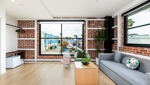 Ein modernes Wohnzimmer mit einem grauen Sofa, einem Holztisch und einem großen Fenster mit Blick auf ein blaues Gebäude und Palmen.