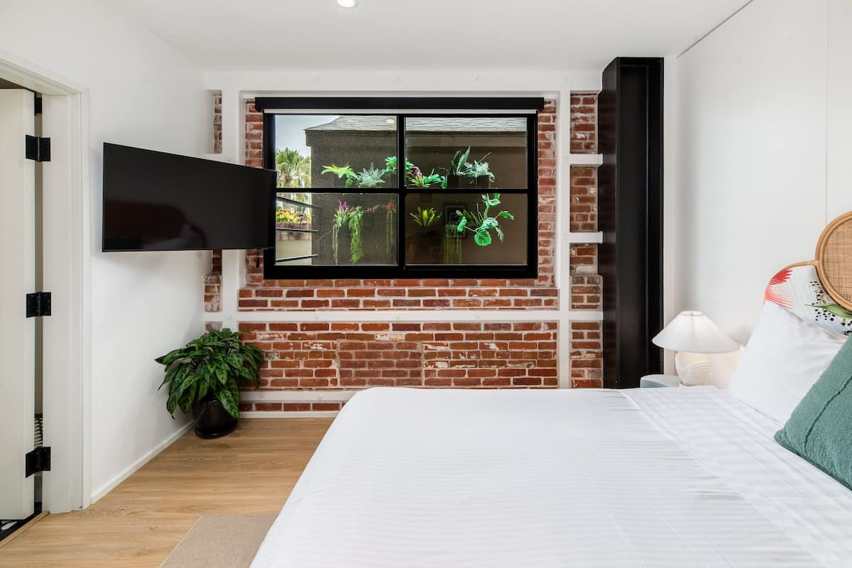A bedroom with a brick wall, a large window with plants, a bed, and a TV mounted on the wall.