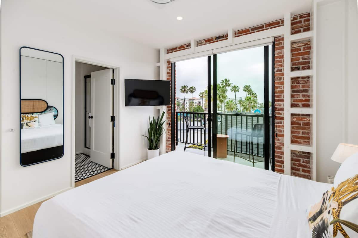 A modern bedroom with a large bed, a TV mounted on the wall, and a balcony with a view of palm trees.