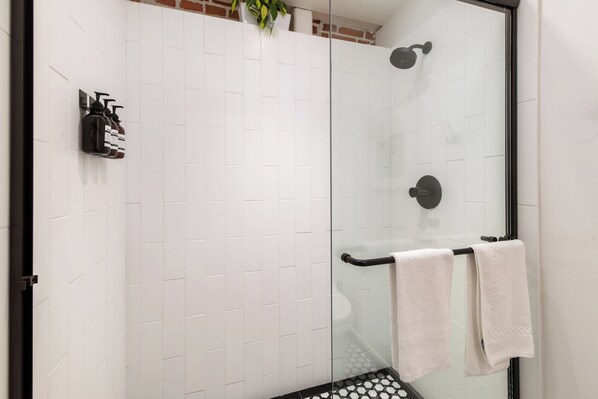 A glass-enclosed shower with white tiled walls, a black towel rack, and a potted plant. - The Brick Hotel (Oceanside)