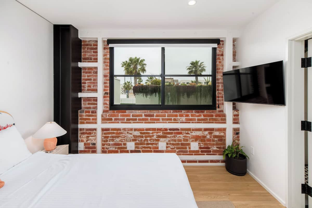 A modern bedroom with a brick wall, a large window, a bed, a TV, and a potted plant.