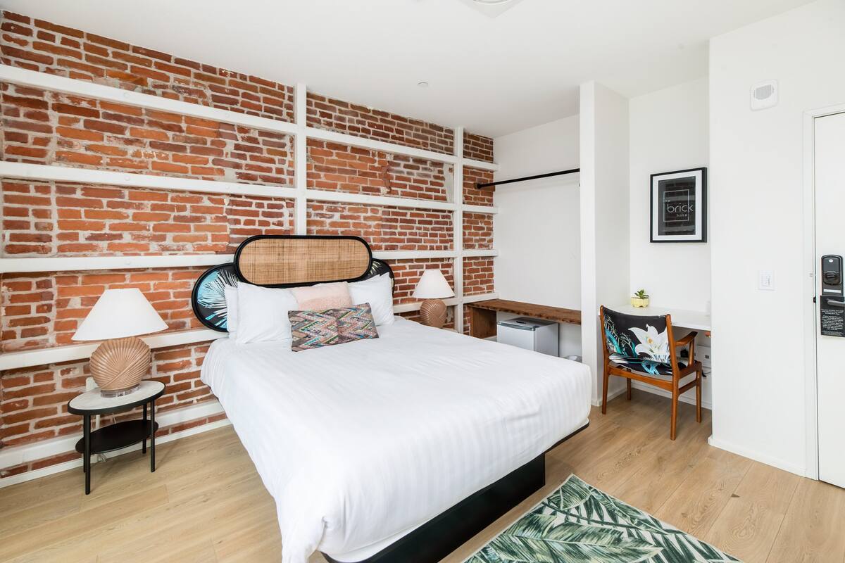 A bedroom with a brick wall, a bed with white bedding, two bedside tables with lamps, a small desk, and a chair.
