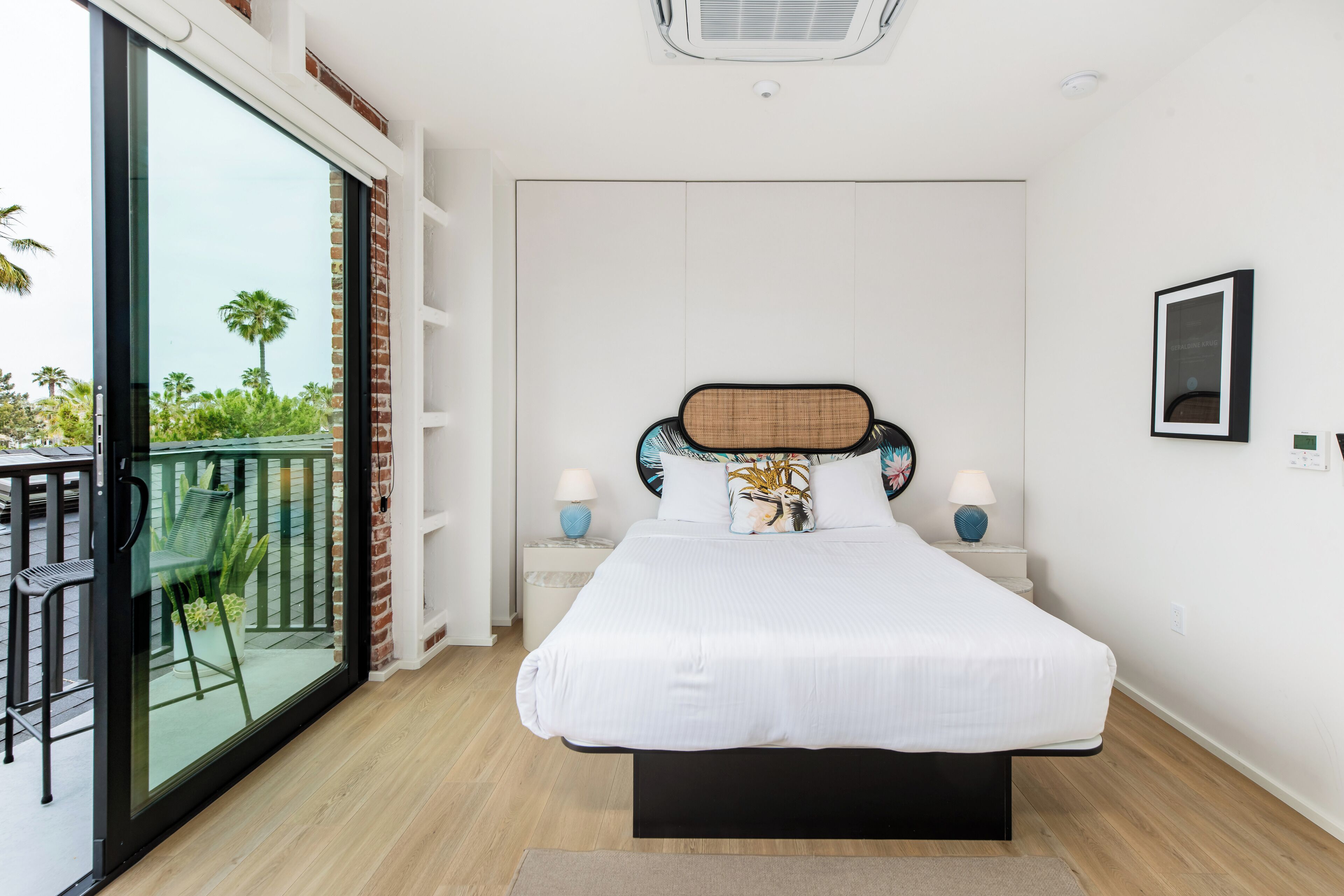 A bedroom with a large bed, two bedside tables with lamps, a glass door leading to a balcony with chairs, and a view of palm trees.