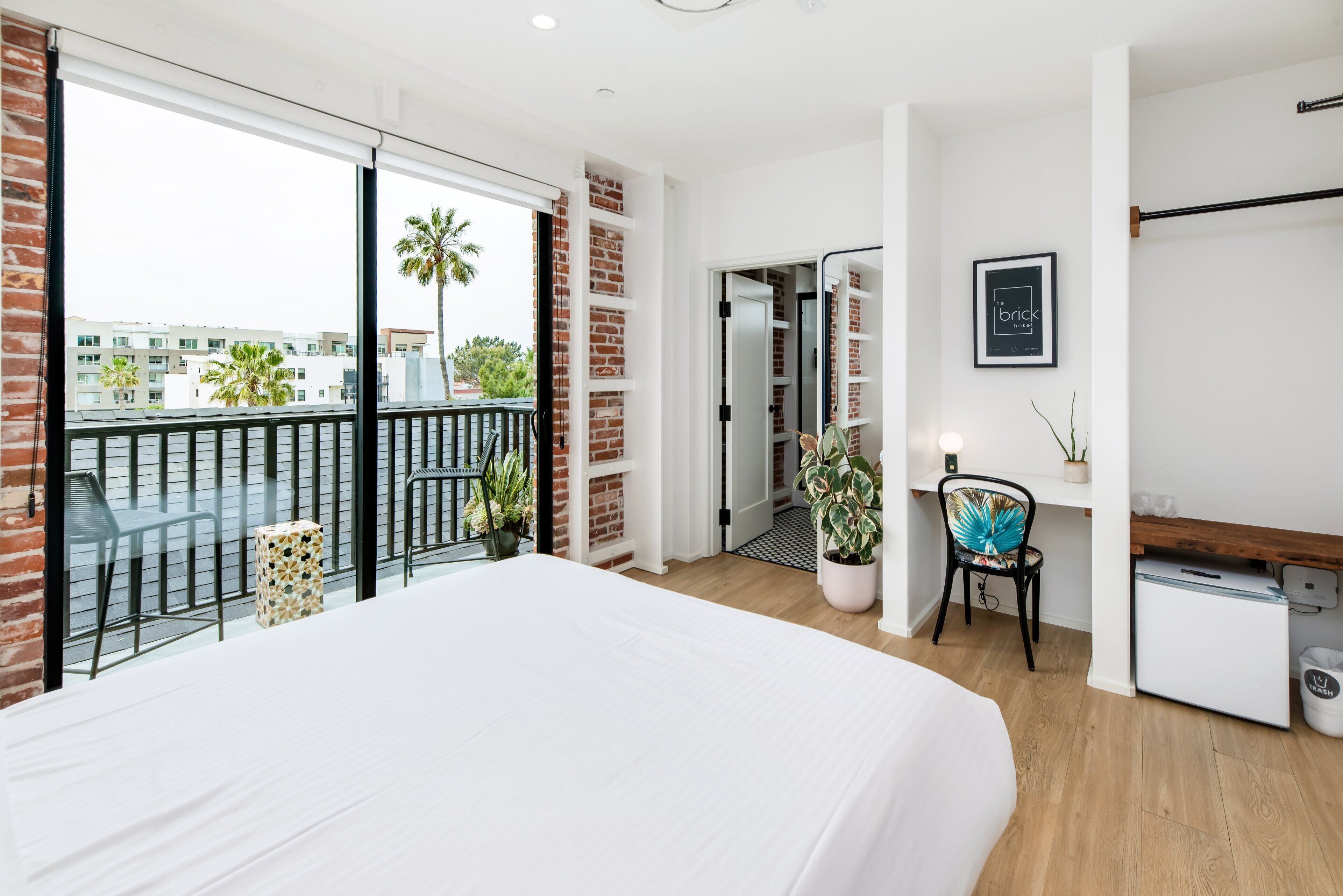 A modern hotel room with a large bed, a balcony with a view of palm trees, a small desk, and a potted plant.