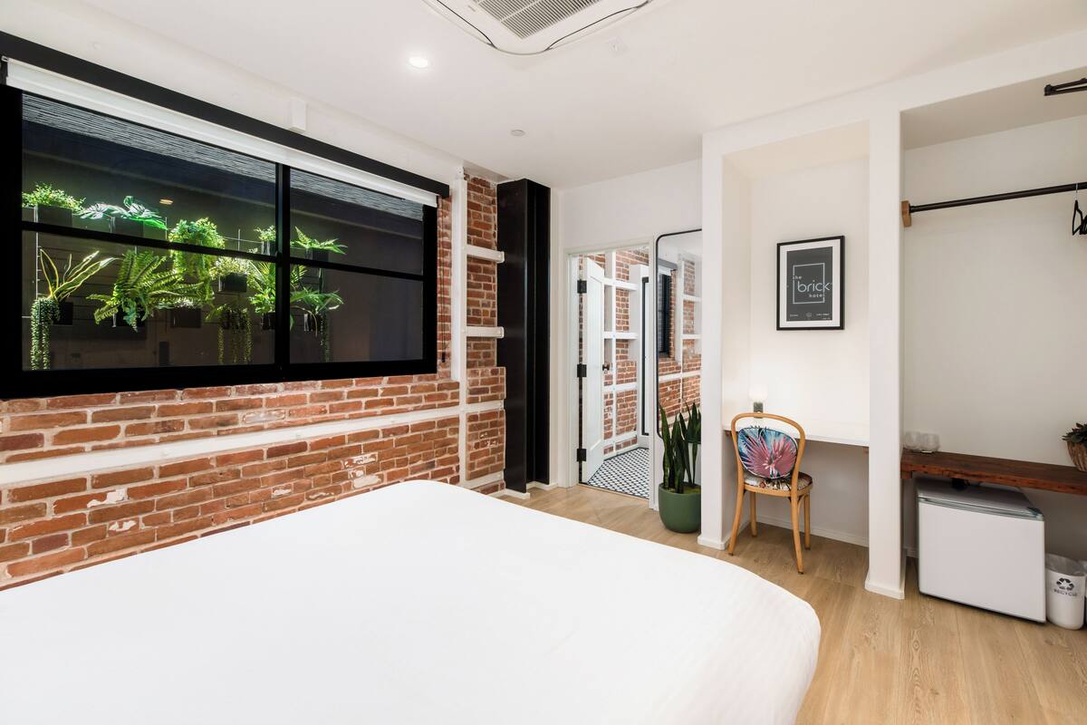 A modern bedroom with a brick wall, a large window with plants, a bed, a desk, and a small refrigerator.