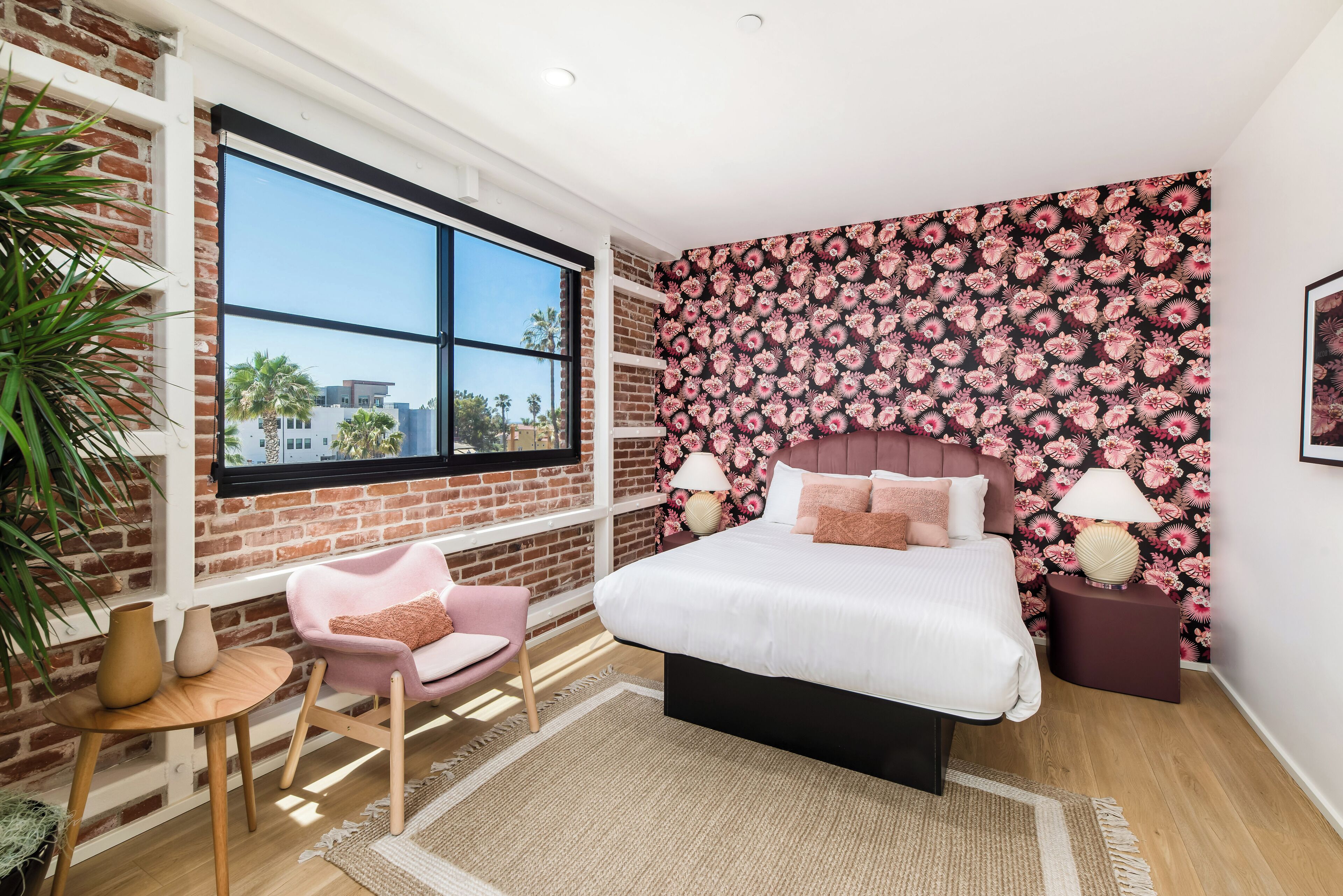 A bedroom with a large window, a brick wall, a floral wallpaper, a bed with white bedding, a pink chair, and a small table with vases.