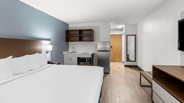 A modern hotel room with a large bed, a kitchenette with a microwave, refrigerator, and wooden cabinets, a flat-screen TV mounted on the wall, and a wooden door leading to another room. - Suburban Studios Chester I-95, VA (Chester)