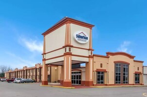 Un hotel para estancias prolongadas en una zona residencial, con entrada central, varios espacios de estacionamiento y un cielo azul despejado.