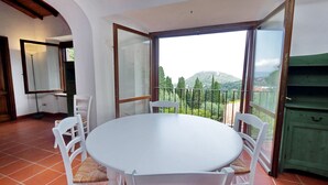 Ein Essbereich mit einem runden Tisch und weißen Stühlen, großen Fenstern mit Blick auf Grünflächen und Hügel, sowie einem grünen Schrank.