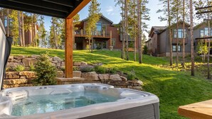Un bain à remous dans un jardin, avec un mur en pierre, une terrasse en bois et une vue sur une maison à plusieurs étages dotée d’un balcon.