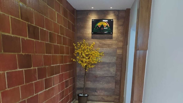 Un pasillo con pared de ladrillo, pared de madera con un letrero, un árbol amarillo en maceta y dos flores rojas en macetas.