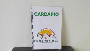 Una libreta de espiral con el título "CARDÁPIO" y el logotipo de "HOSTEL VILA BELA" sobre una superficie negra reflectante.