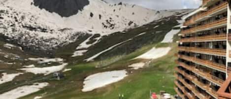 Bergachtig landschap met een opvallende bergtop, een gebouw met meerdere balkons, en sneeuwvlekken.