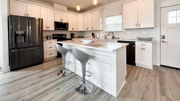 A modern kitchen with white cabinets, stainless steel appliances, and a central island with bar stools. - Beach Cottage with a View!! (Gulfport)